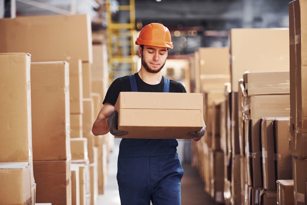 Young warehouse worker in uniform and hard hat carries box in hands.